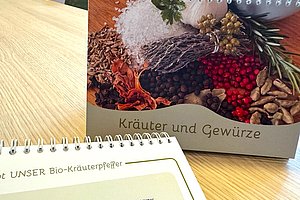 Zwei auf einem Holztisch stehende Spiralblockkalender, einer mit dem Titel 'Kräuter und Gewürze' und einem Bild verschiedener Gewürze, der andere mit Text und einer Überschrift zu Bio-Kräuterpfeffer.