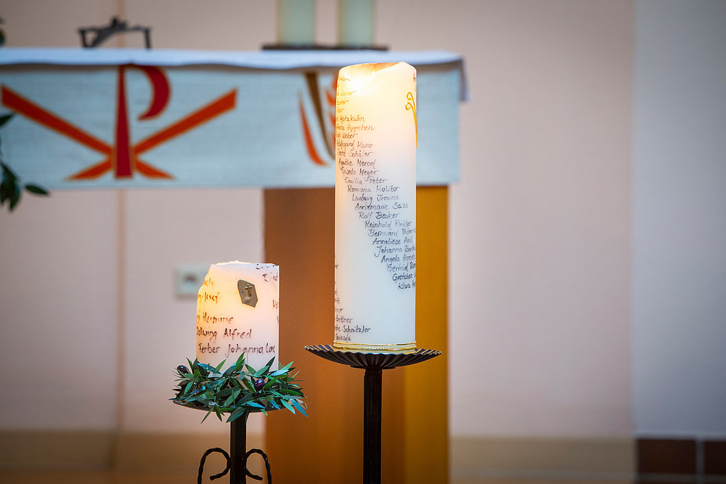 Zwei brennende Kerzen auf Metallständern, eine mit handgeschriebenen Namen, im Hintergrund ein Altar mit christlichem Symbol