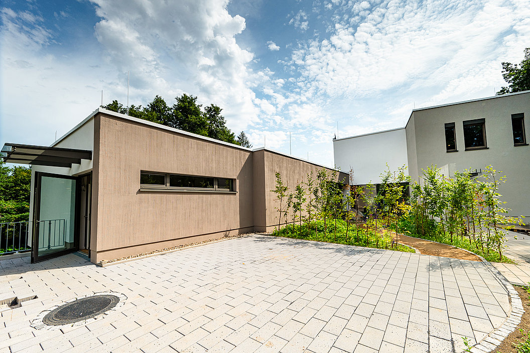 Modernes, eingeschossiges Gebäude mit flachem Dach, hellbrauner Fassade und großen Fenstern, umgeben von gepflastertem Hof und Büschen.