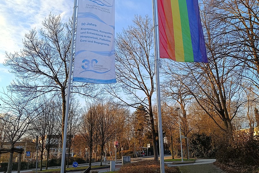Zwei Fahnen an Masten vor kahlen Bäumen, eine mit Regenbogenfarben, die andere mit Text und Logo in Blau auf Weiß.