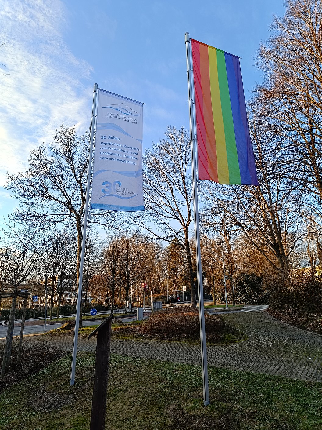 Zwei Fahnen an Masten vor kahlen Bäumen, eine mit Regenbogenfarben, die andere mit Text und Logo in Blau auf Weiß.