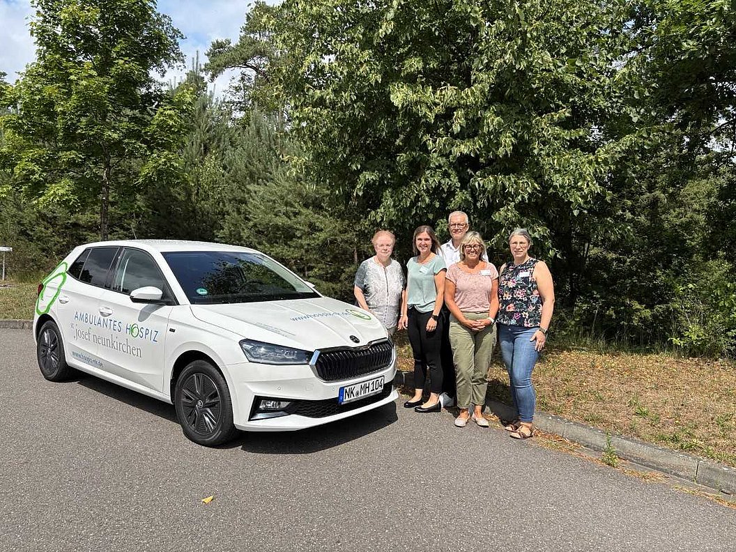 Weißes Auto mit der Aufschrift 'AKB Diakonie Hospiz' steht auf einer Straße neben fünf Personen vor Bäumen.