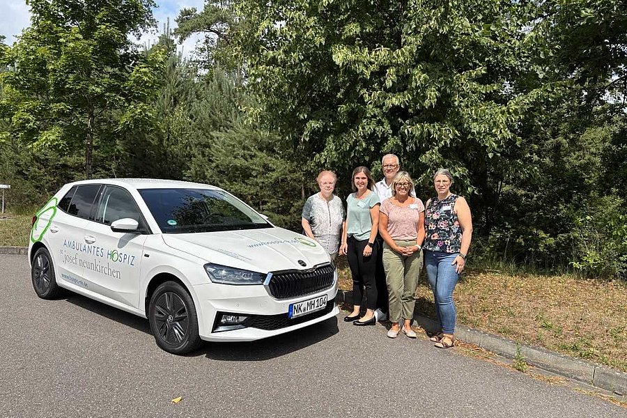 Weißes Auto mit der Aufschrift 'AKB Diakonie Hospiz' steht auf einer Straße neben fünf Personen vor Bäumen.