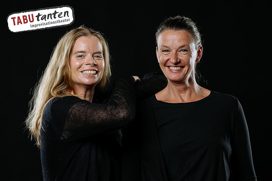Zwei Frauen in schwarzer Kleidung vor schwarzem Hintergrund, eine mit langen blonden Haaren, die andere mit zurückgebundenem Haar. Logo 'TABUtanten Improvisationstheater' oben links.
