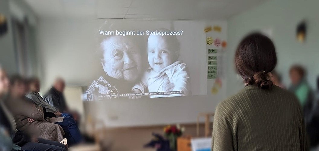 Person betrachtet eine Projektion mit einer alten Frau und einem Baby, darüber der Text 'Wann beginnt der Sterbeprozess?' in einem Raum mit mehreren sitzenden Personen