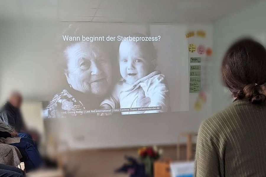Person betrachtet eine Projektion mit einer alten Frau und einem Baby, darüber der Text 'Wann beginnt der Sterbeprozess?' in einem Raum mit mehreren sitzenden Personen