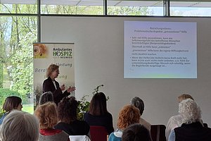 Frau hält Vortrag vor Publikum in hellem Raum mit großer Fensterfront und projiziertem Text zur Begleitung von Hospizarbeit
