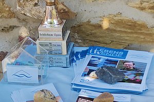 Silberner Pokal mit der Aufschrift 'Schlickum Open' neben mehreren Flyern und Broschüren, auf denen Steine liegen, auf einem weißen Tuch vor einer Steinmauer.