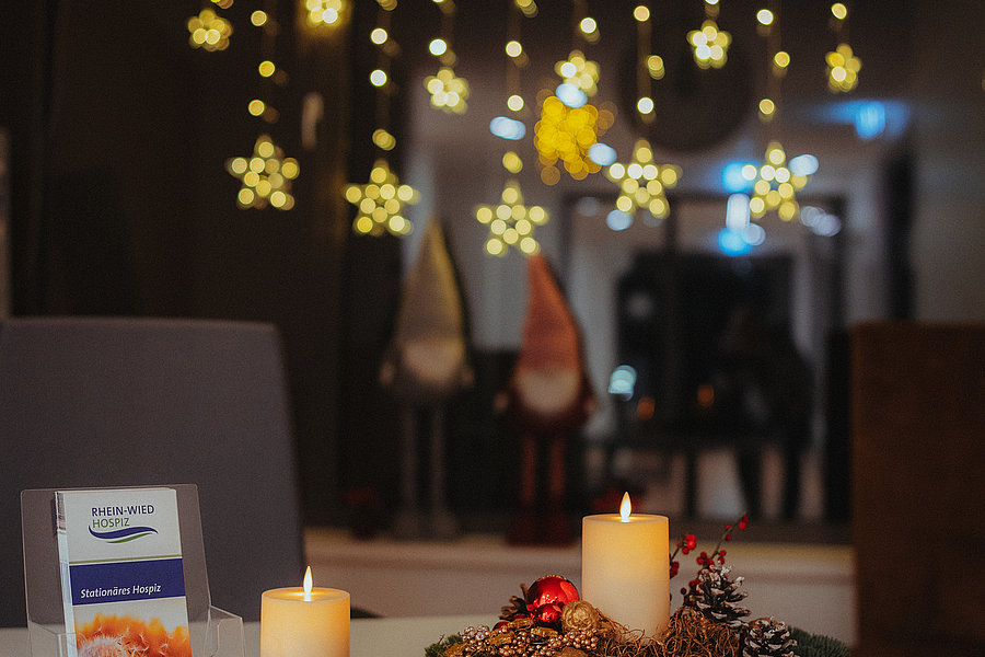 Weihnachtliche Tischdekoration mit zwei brennenden Kerzen, Tannenzweigen, Zapfen und roten Kugeln, im Hintergrund leuchtende Sternlichter.