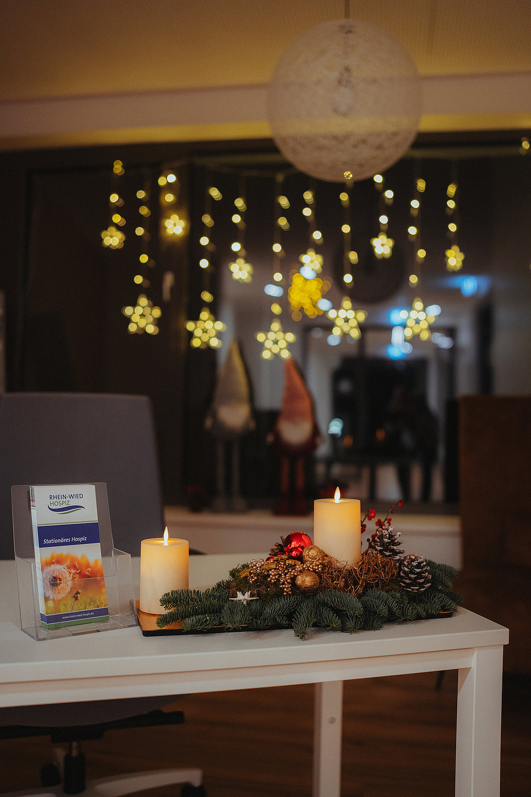 Weihnachtliche Tischdekoration mit zwei brennenden Kerzen, Tannenzweigen, Zapfen und roten Kugeln, im Hintergrund leuchtende Sternlichter.