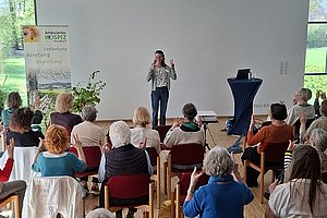 Frau steht vor Leinwand und spricht vor Publikum in hellem Raum mit großen Fenstern und Banner links