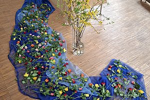 Blaue Stoffbahn auf Holzboden mit darauf liegenden Rosen und daneben stehender Glasvase mit gelb blühenden Zweigen