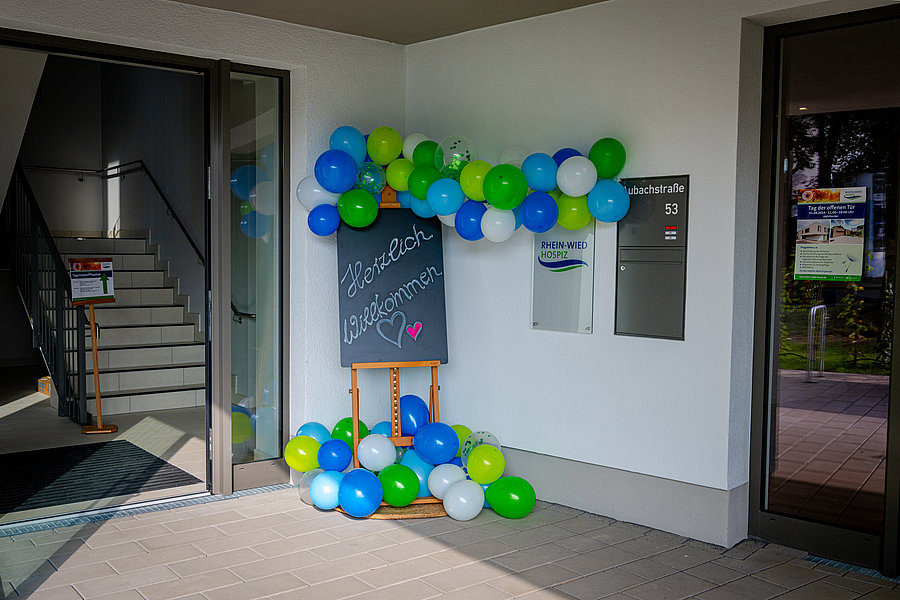Eingangsbereich mit geöffneten Glastüren, bunte Luftballons in Blau, Grün und Weiß, Tafel mit der Aufschrift 'Herzlich willkommen' und einem Herzsymbol.