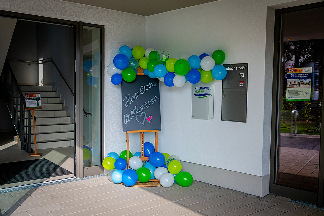 Eingangsbereich mit geöffneten Glastüren, bunte Luftballons in Blau, Grün und Weiß, Tafel mit der Aufschrift 'Herzlich willkommen' und einem Herzsymbol.