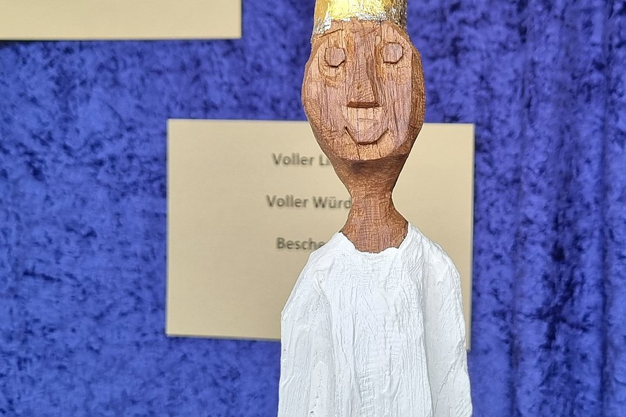 Holzfigur mit goldener Krone und weißem Gewand vor blauem Hintergrund, im Hintergrund zwei beige Zettel mit Text.
