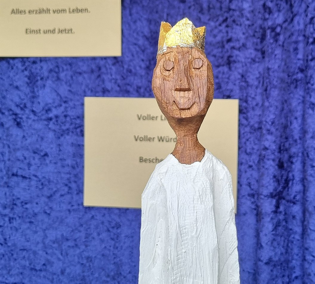 Holzfigur mit goldener Krone und weißem Gewand vor blauem Hintergrund, im Hintergrund zwei beige Zettel mit Text.