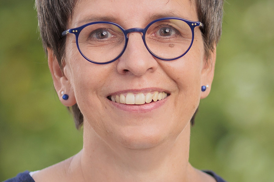 Hiltrud Sachs mit kurzem, grauem Haar trägt eine dunkelblaue Strickjacke über einem weißen Oberteil vor unscharf grünem Hintergrund.
