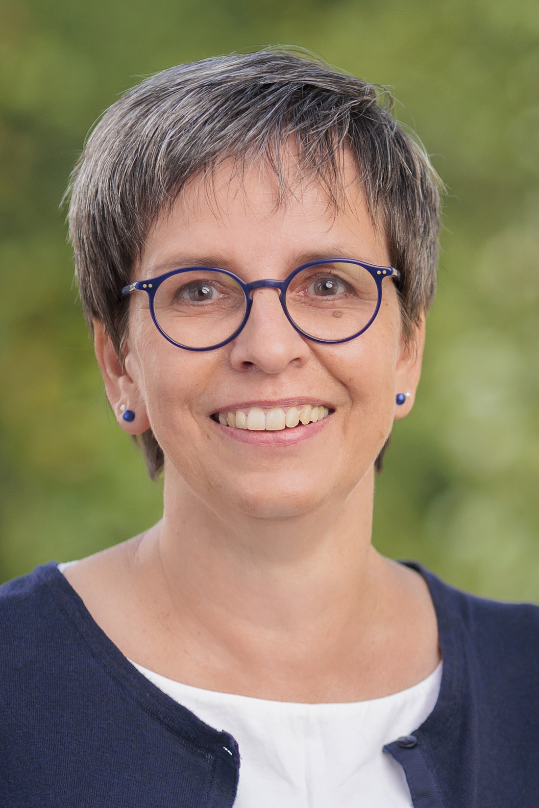Hiltrud Sachs mit kurzem, grauem Haar trägt eine dunkelblaue Strickjacke über einem weißen Oberteil vor unscharf grünem Hintergrund.