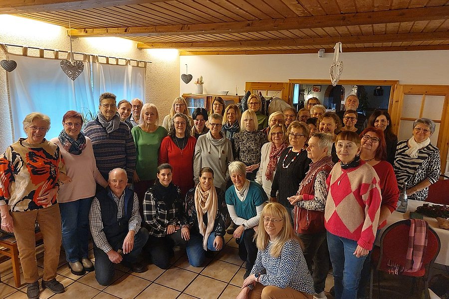 Gruppenfoto von 24 Personen in einem Raum mit Holzbalkendecke und Kachelfußboden, einige sitzen, andere stehen, Kleidung in verschiedenen Farben und Mustern.