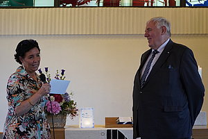 Frau mit gemustertem Kleid hält Mikrofon und Zettel, steht vor Blumengesteck, Mann im dunklen Anzug mit Krawatte steht ihr gegenüber.