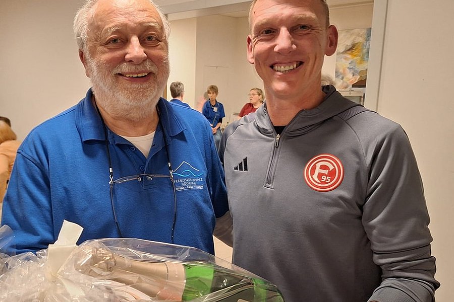 Zwei Männer stehen nebeneinander, einer trägt blaues Hemd mit Brille am Kragen, der andere graues Adidas-Sportshirt mit Fortuna-Düsseldorf-Logo, sie halten eine Geschenkverpackung mit Flasche und Buch.