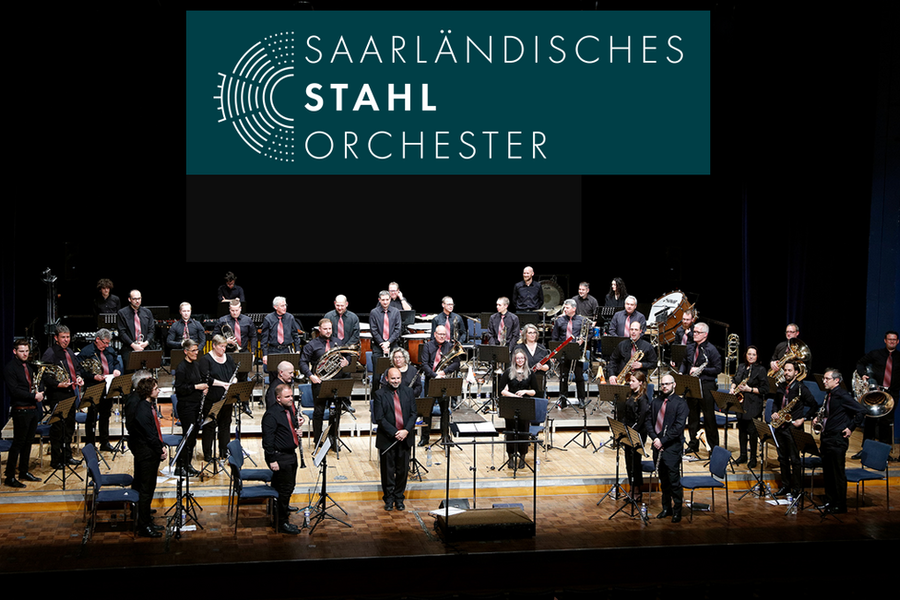 Orchester auf Bühne mit verschiedenen Blasinstrumenten und Streichinstrumenten, im Hintergrund Banner mit Schrift 'Saarländisches Stahl Orchester'.
