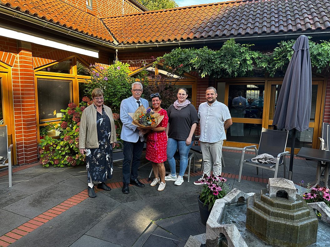 Fünf Personen stehen auf einem gepflasterten Hof vor einem Backsteingebäude, eine Person hält einen Blumenstrauß.