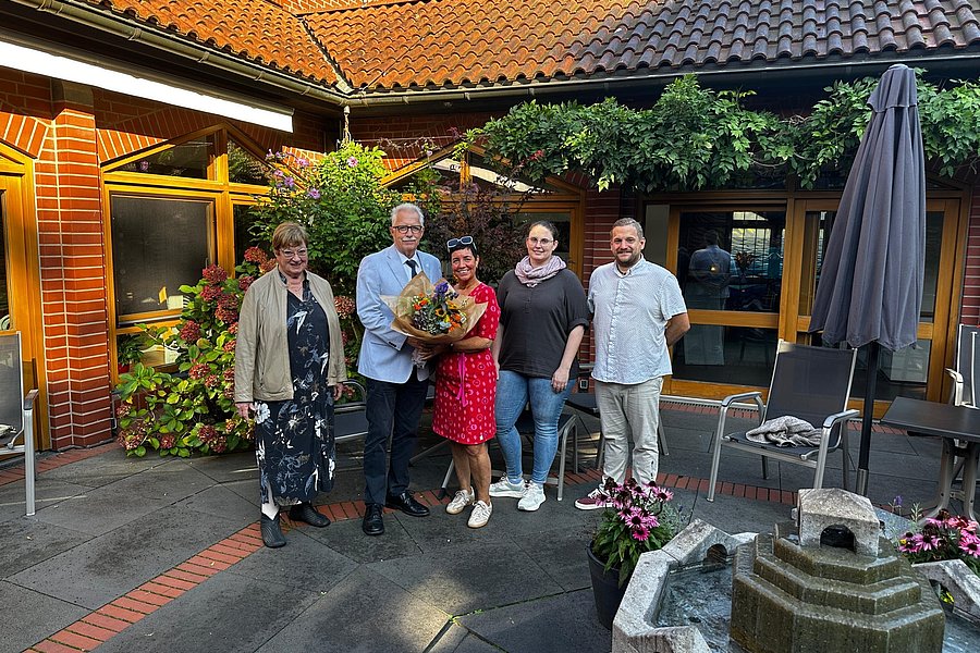 Fünf Personen stehen auf einem gepflasterten Hof vor einem Backsteingebäude, eine Person hält einen Blumenstrauß.