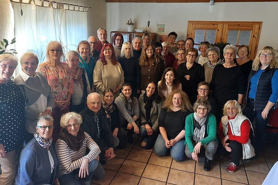 Gruppenfoto von 27 Personen in einem Raum mit Kacheln, Vorhängen und Holzmöbeln, einige sitzen, andere stehen, verschiedene Kleidung und Frisuren sichtbar.