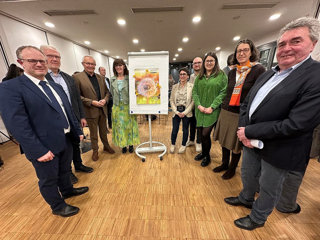 Gruppe von Menschen steht im Halbkreis um ein Flipchart mit einem Bild einer Sonnenblume in einem Raum mit Holzfußboden und Deckenleuchten.