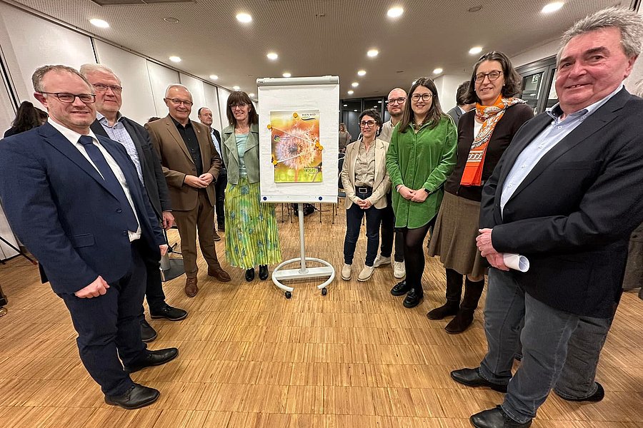Gruppe von Menschen steht im Halbkreis um ein Flipchart mit einem Bild einer Sonnenblume in einem Raum mit Holzfußboden und Deckenleuchten.