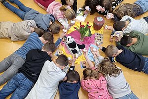 Kinder liegen im Kreis auf dem Boden und greifen nach Spielmaterial in der Mitte