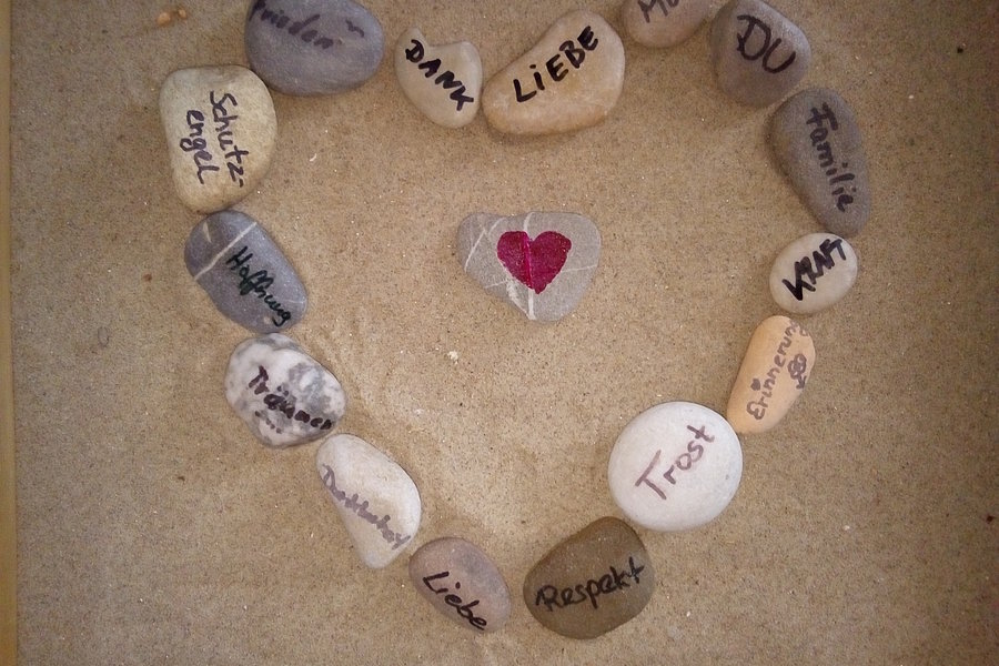 Herzform aus Steinen auf Sand, in der Mitte ein Stein mit rotem Herz, umgeben von Steinen mit deutschen Worten wie 'Danke', 'Liebe', 'Mut', 'Familie', 'Respekt' und 'Vertrauen'.