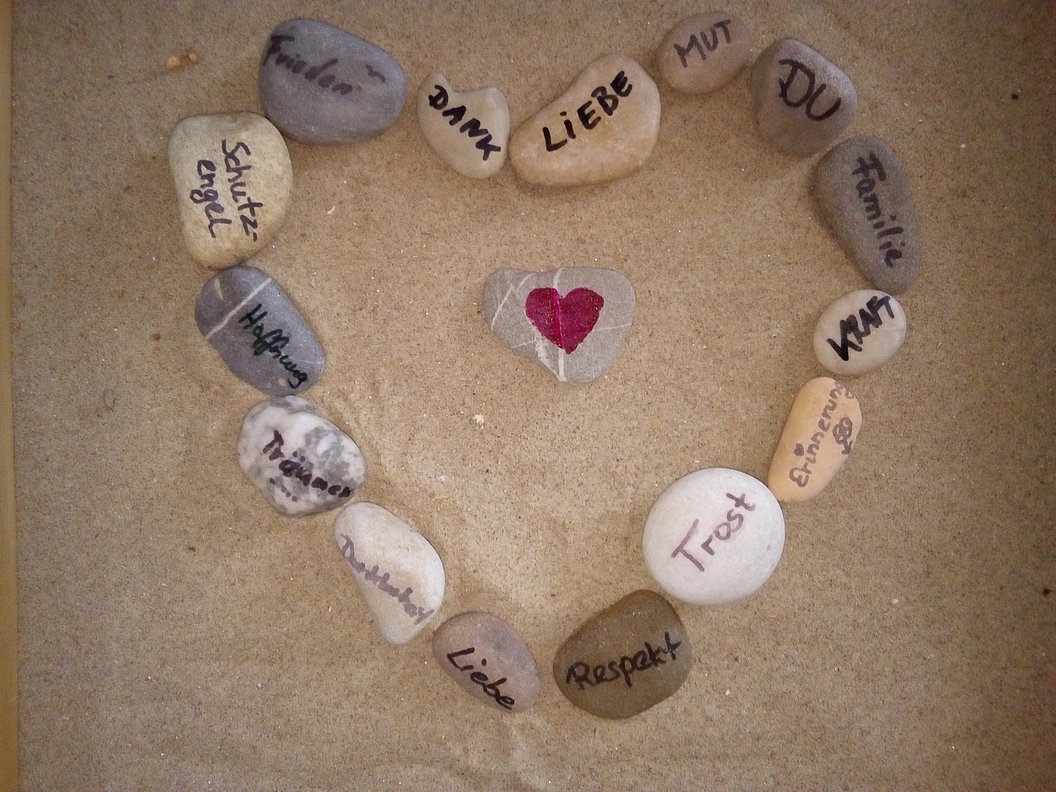 Herzform aus Steinen auf Sand, in der Mitte ein Stein mit rotem Herz, umgeben von Steinen mit deutschen Worten wie 'Danke', 'Liebe', 'Mut', 'Familie', 'Respekt' und 'Vertrauen'.