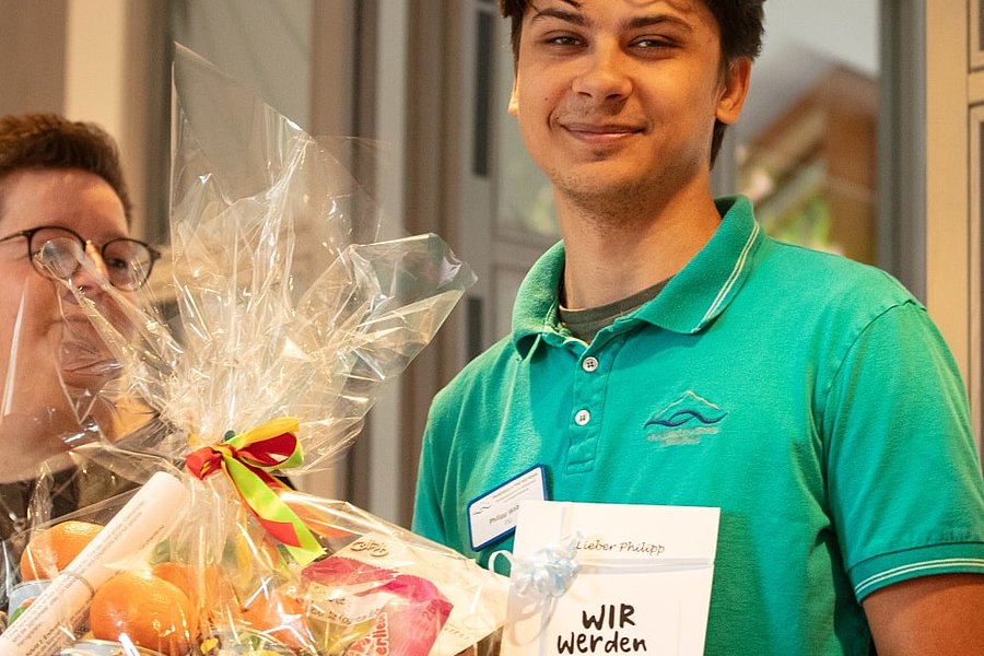 Mann in grünem Poloshirt hält einen Geschenkkorb mit bunten Lebensmitteln und eine Karte mit der Aufschrift 'WIR WERDEN DIR FEHLEN'.