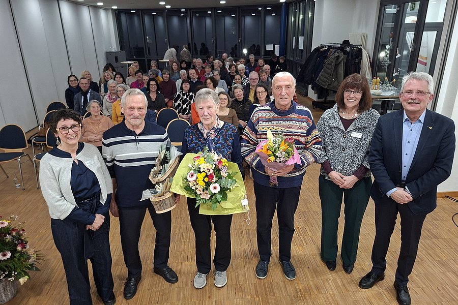 Feierliche Veranstaltung mit mehreren Personen, Blumensträußen und einer großen Gruppe im Hintergrund.
