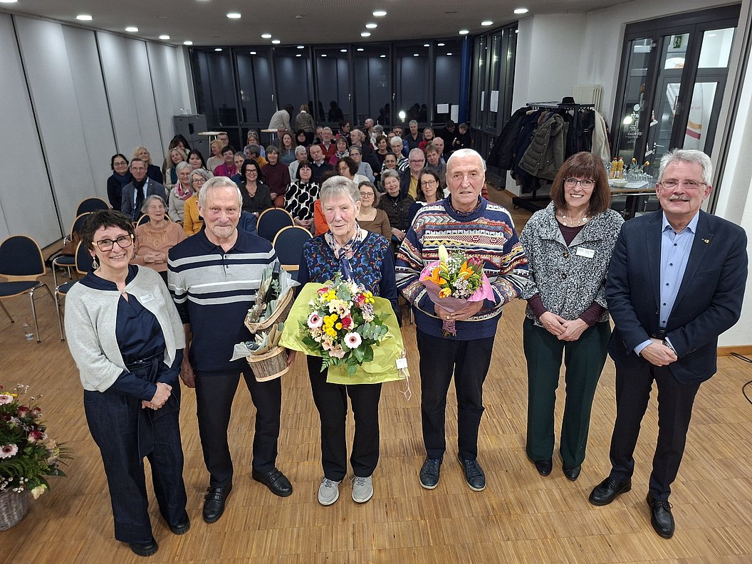 Feierliche Veranstaltung mit mehreren Personen, Blumensträußen und einer großen Gruppe im Hintergrund.