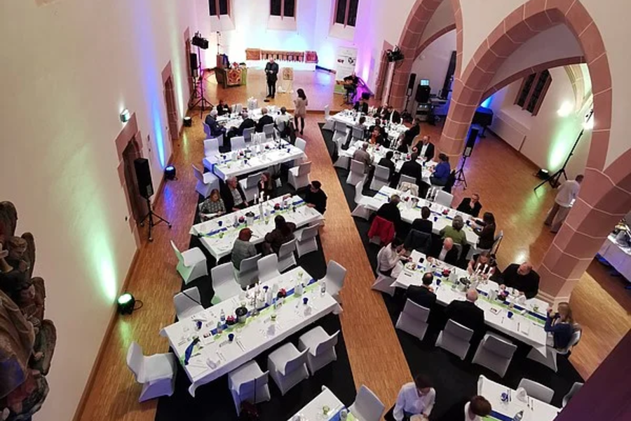 Blick von oben auf einen festlich gedeckten Saal mit mehreren langen Tischen und weißen Stühlen in einer Kirche mit hohen, spitzbogigen Fenstern und Gewölbedecke.