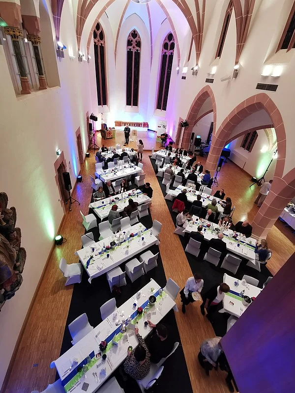 Blick von oben auf einen festlich gedeckten Saal mit mehreren langen Tischen und weißen Stühlen in einer Kirche mit hohen, spitzbogigen Fenstern und Gewölbedecke.
