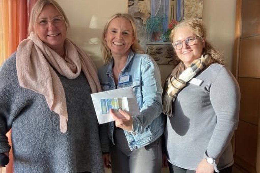 Drei Frauen stehen nebeneinander in einem Raum, eine hält eine Postkarte mit einem blauen Motiv, im Hintergrund ein Bild mit einer blauen Tür.