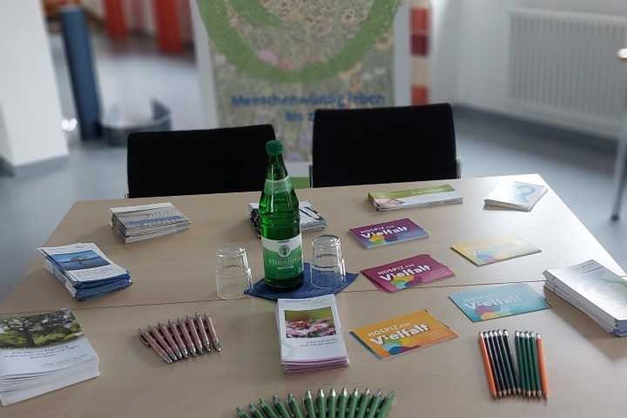 Tisch mit grünen Kugelschreibern, Flyern und einer Wasserflasche vor einem Banner des Ambulanten Hospiz Josef Neunkirchen in einem hellen Raum mit Fenstern.