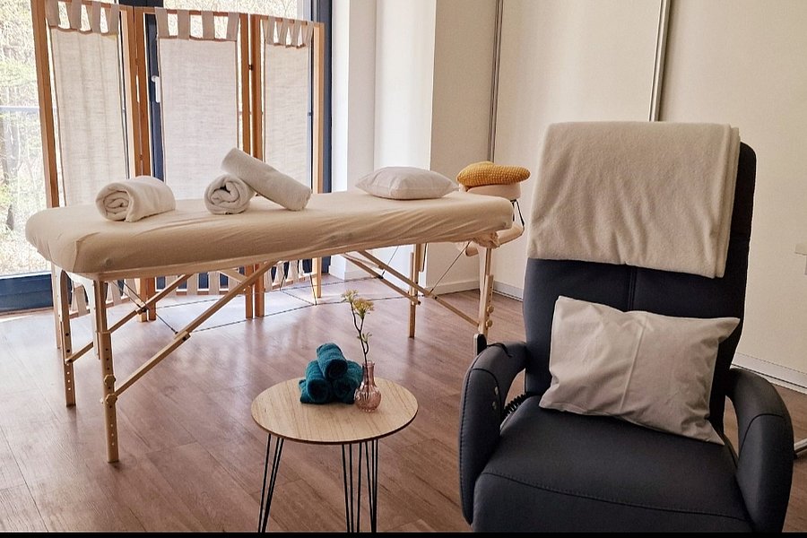 Massageliege mit weißen Handtüchern und Kissen in hellem Raum, daneben schwarzer Sessel mit Kissen und Decke, kleiner runder Tisch mit blauer Rolle und Vase.