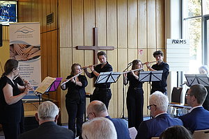 Fünf Musiker spielen Querflöte vor einem Holzwand mit Kreuz, Publikum sitzt im Vordergrund.