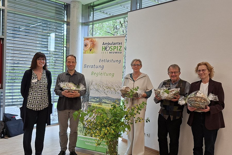 Fünf Personen stehen in einem Raum vor einem Banner mit dem Schriftzug 'Ambulantes Hospiz' und halten jeweils ein Geschenk oder Blumen
