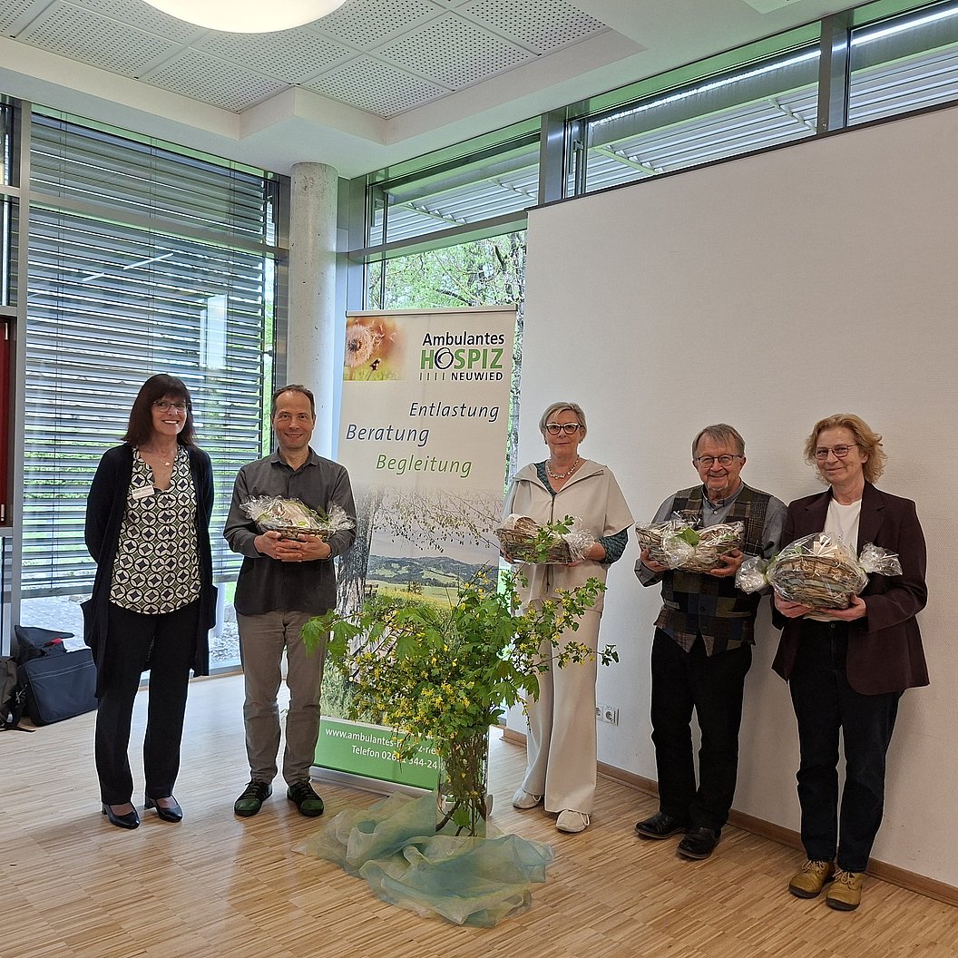 Fünf Personen stehen in einem Raum vor einem Banner mit dem Schriftzug 'Ambulantes Hospiz' und halten jeweils ein Geschenk oder Blumen