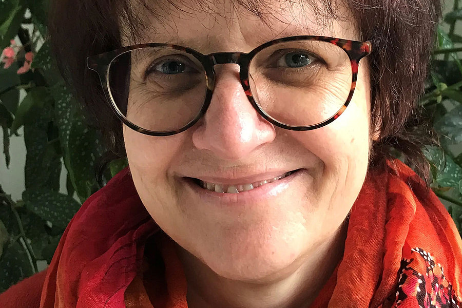 Dr. Karin Rodrian mit kurzen braunen Haaren trägt eine orange-rote Schal mit schwarzem und rotem Muster vor grünem Pflanzenhintergrund.