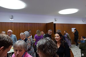 Mehrere Personen in einem Raum mit Holzvertäfelung an der Wand und zwei runden Deckenleuchten, die sich unterhalten und stehen.