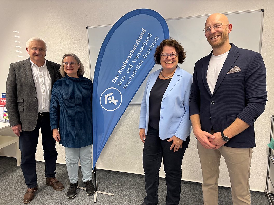 Vier Personen stehen vor einem blauen Werbefahnen mit der Aufschrift 'Der Kinderschutzbund Orts- und Kreisverband Neuss und Bad Dürkheim' in einem Raum mit weißer Wand und Möbeln