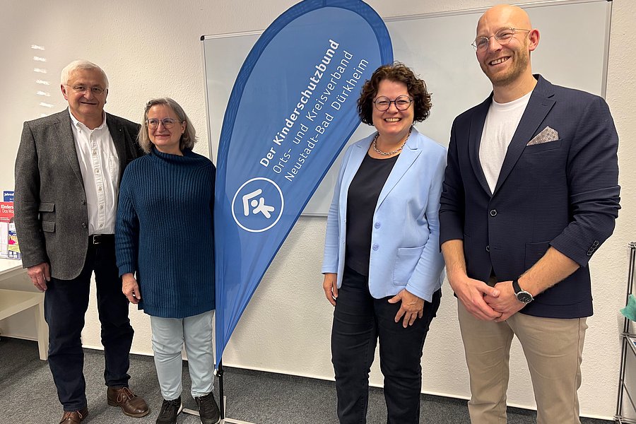 Vier Personen stehen vor einem blauen Werbefahnen mit der Aufschrift 'Der Kinderschutzbund Orts- und Kreisverband Neuss und Bad Dürkheim' in einem Raum mit weißer Wand und Möbeln