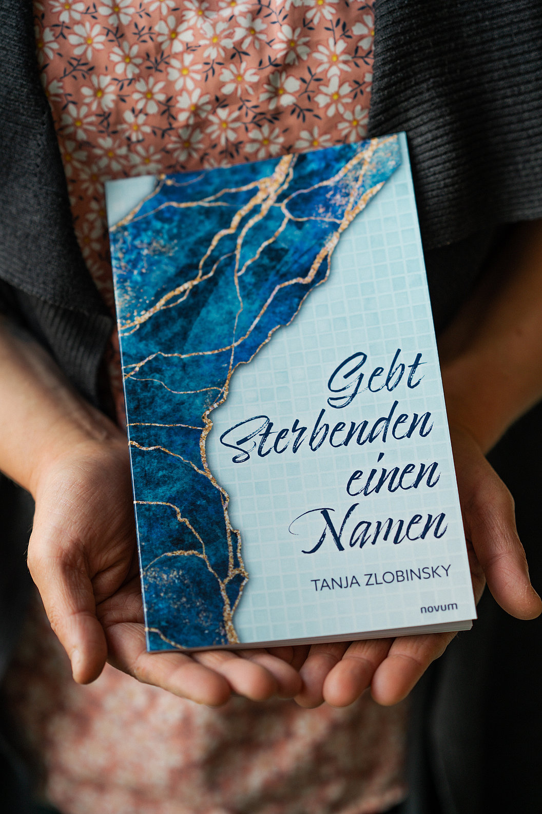 Hände halten ein Buch mit blau-weißem Marmormuster und dem Titel 'Gebt Sterbenden einen Namen' von Tanja Zlobinsky